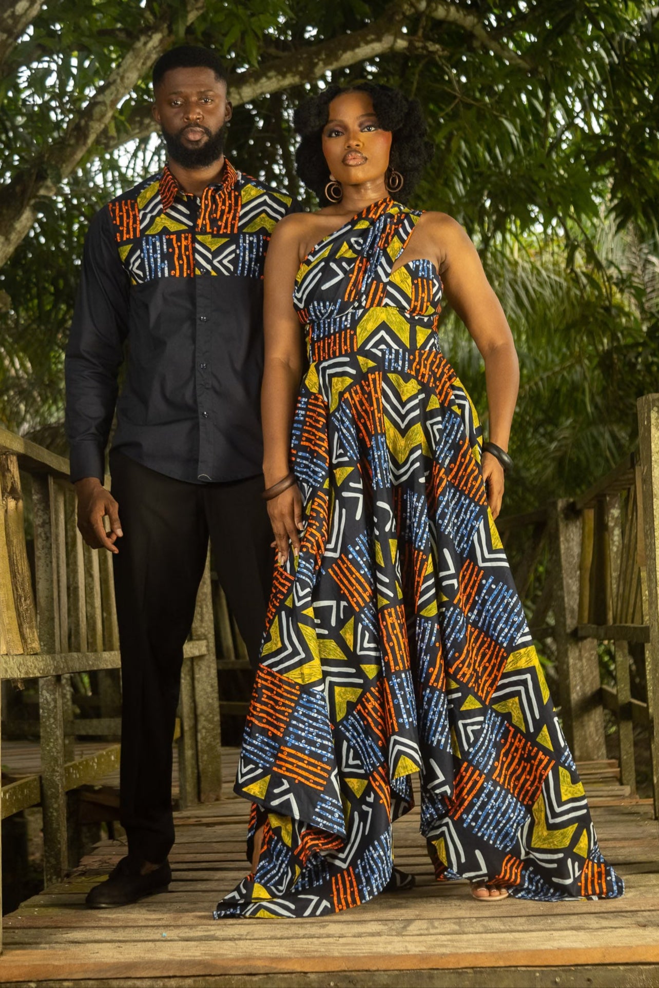 Two people in traditional African attire standing on a wooden bridge in a forest setting