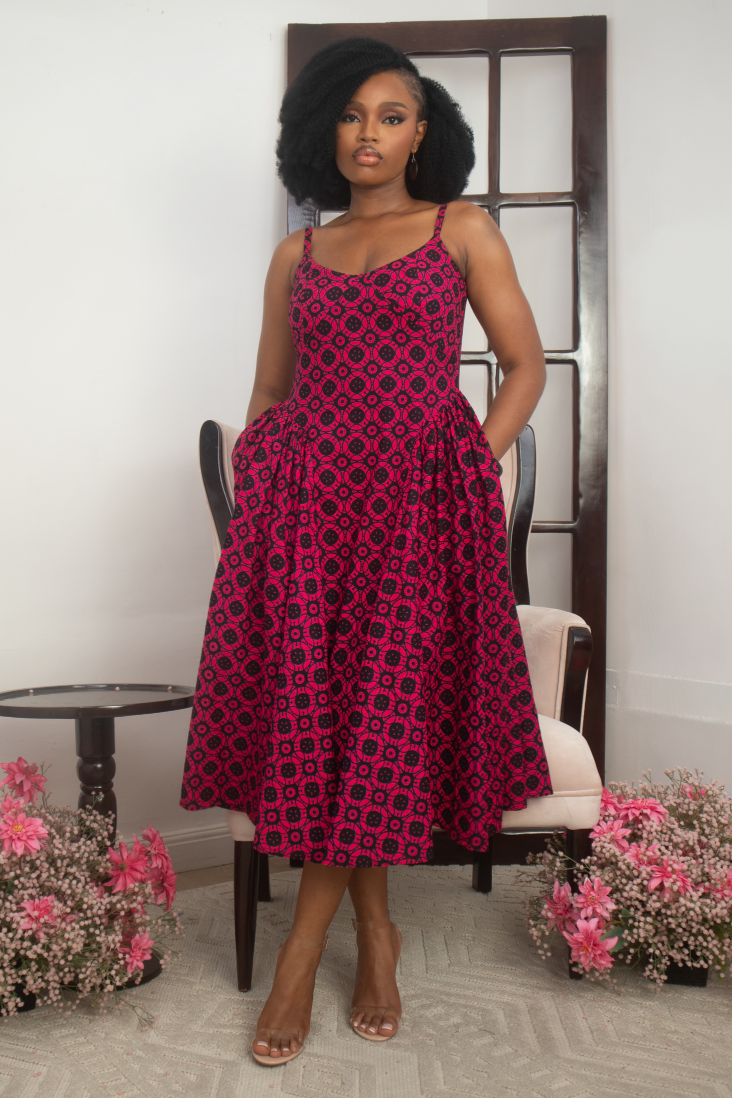 Woman wearing a pink and black patterned African dress standing in a room with flowers and a chair.